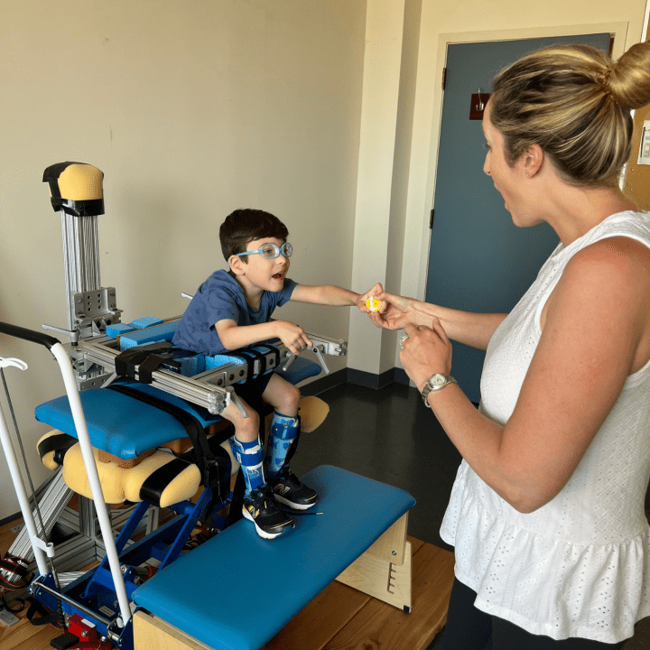 A boy wearing blue glasses and a blue shirt is sitting on a bench with supports around his waste while he is reaching for a toy from a physical therapist.