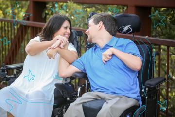 Shelby, a dark haired woman wearing a white dress sitting in a wheelchair next to David who is wear in Blue shirt and khaki pants sitting in his wheelchair.