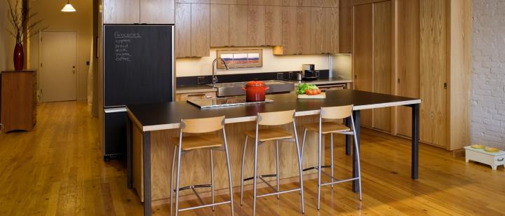 Photo of modern kitchen with wooden paneling. Island bench with 3 stools and black bench top
