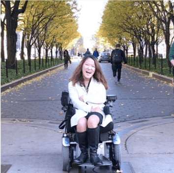 Writer Sarah Kim sitting in her power wheelchair in front of an avenue of trees