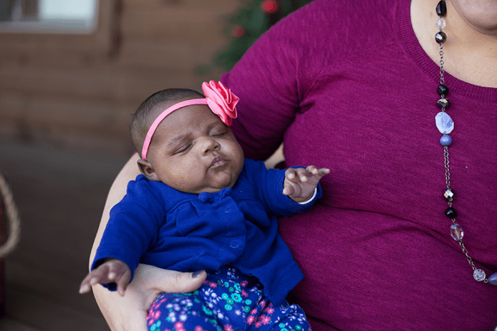 Photo of baby girl who is one month old in a blue sweater with pink flower headband. She is being held in her mother's arms.