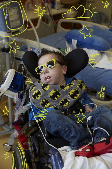 Photo of a young man in a blue shirt, wearing dark sunglasses with yellow rims reclining in a wheelchair with a batman printed neck pillow around his neck. He is in a hospital room and has a feeding tube attached to the wheelchair.