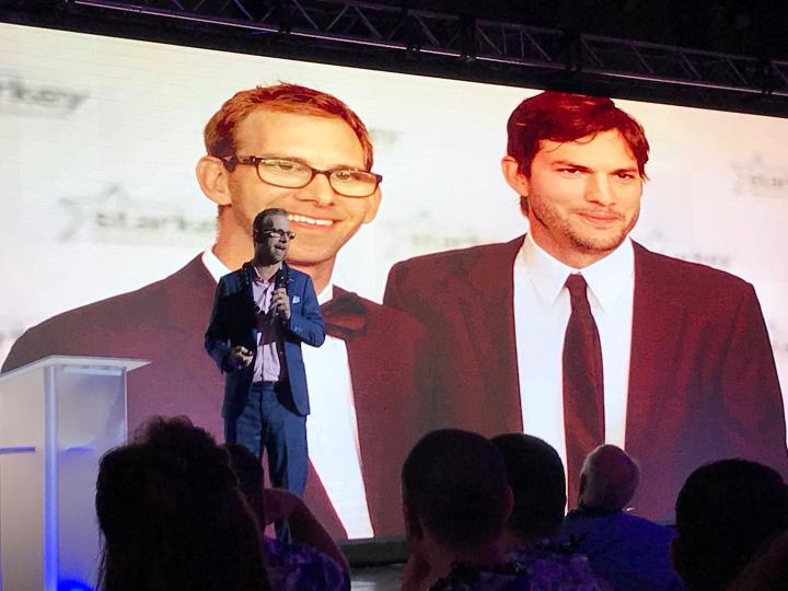 Photo of Michael Kutcher wearing a suit on a stage, holding a microphone. Behind him is a projection of he and ashton kutcher bother wearing black suit and ties