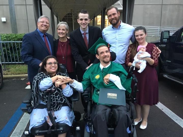 Photograph of a young woman and young man wearing a graduation cap and gown in wheelchairs holding hands with five individuals standing up behind them. One individual is holding an infant.