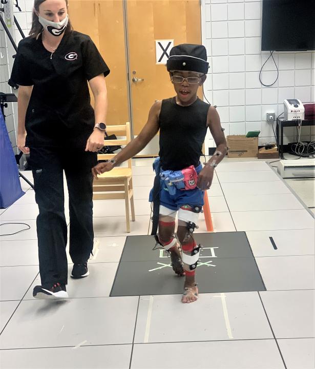 Young boy wearing a helmet on his head and electrodes on his legs walking with a woman wear black scrubs.