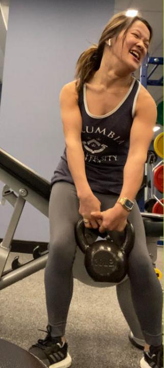 Sarah Kim, standing up holding a black kettle bell while working out, She has long brown hair and is wearing a black tank top with grey leggings and black shoes.