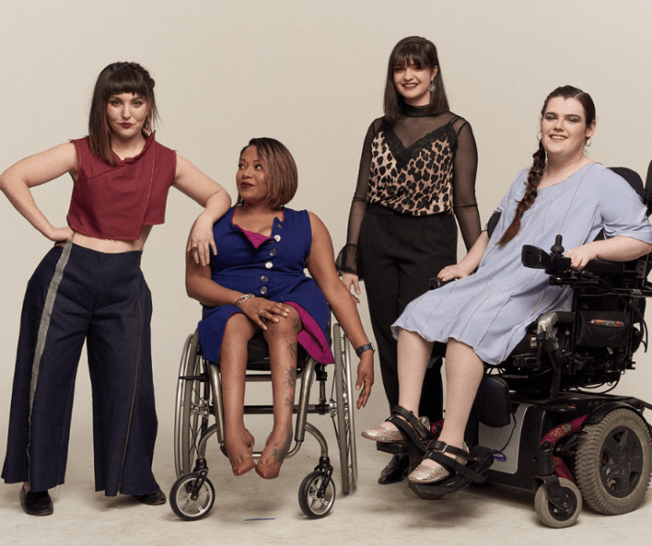 Photo of 4 women: From left to right - White woman standing up with dark shoulder length hair wearing a red sleeveless shirt and black pants. The next woman is seated in a wheelchair. She has dark shoulder length hair and is wearing a blue top. The next woman is standing up with shoulder length dark hair wearing a black and gold top and black pants. The last woman is seated in a powered wheelchair wearing a purple dress. She has a long blonde braid.