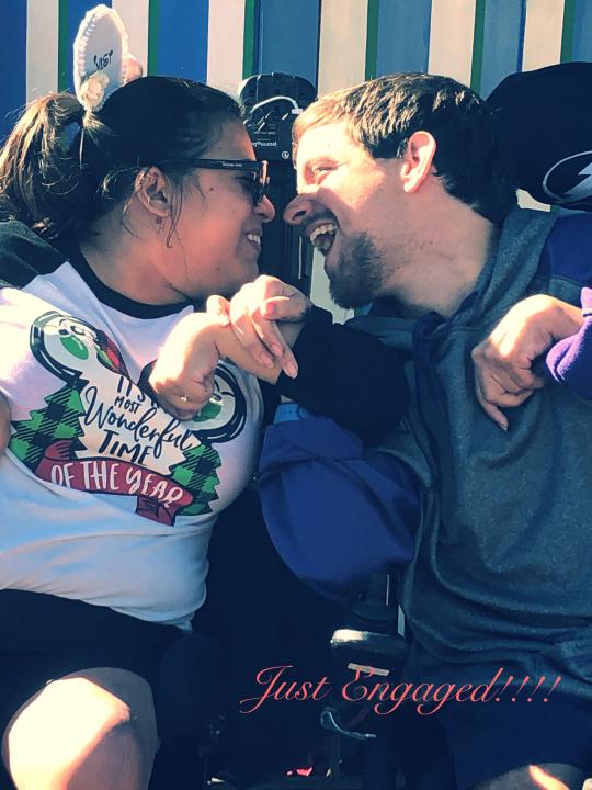 Shelby with long dark hair in a pony tail wear Micky Mouse ears and a white shirt with green lettering holding hands, smiling and looking at David wearing a green shirt. He has brown hair and a short mustache and beard. Both are sitting in their power wheelchairs.