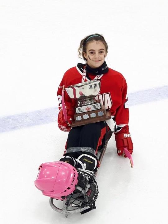 Photo of a young girl with dark hair wearing a red shirt holding a trophy on a sled on the ice rink