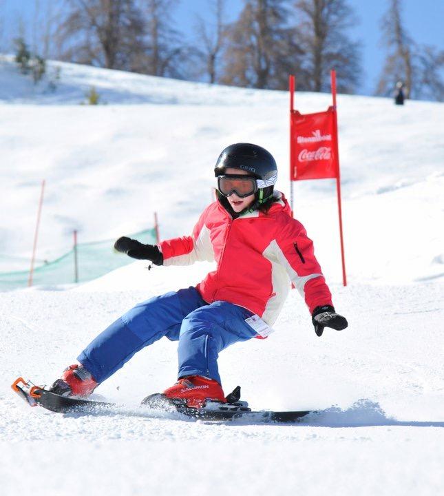 Young person wearing a red and white parka with blue pants and a black helmet snowboarding 