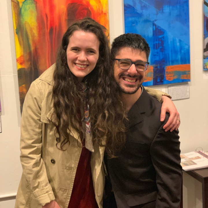 A photo of a young white woman with long brown hair wearing a tan jacket and a red dress with her arm around a young white man with dark hair, glasses and a mustache wearing a black jacket