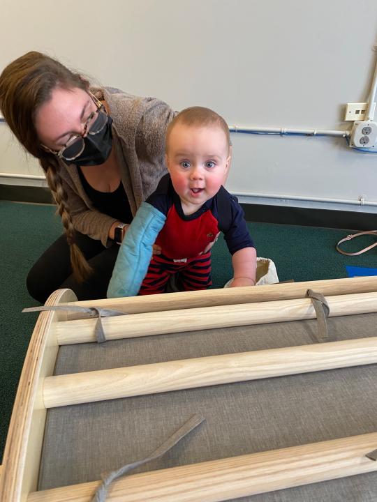 Infant wearing a mitten climbing on a ladder with the help of a caregiver.