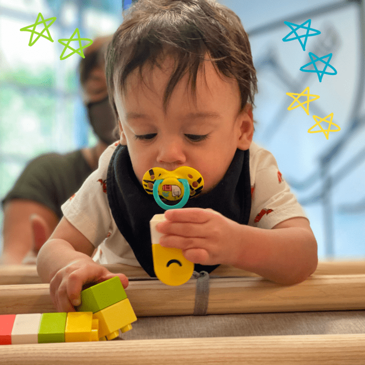 Infant with dark brown hair sucking on a pacifier while playing with blocks on his belly