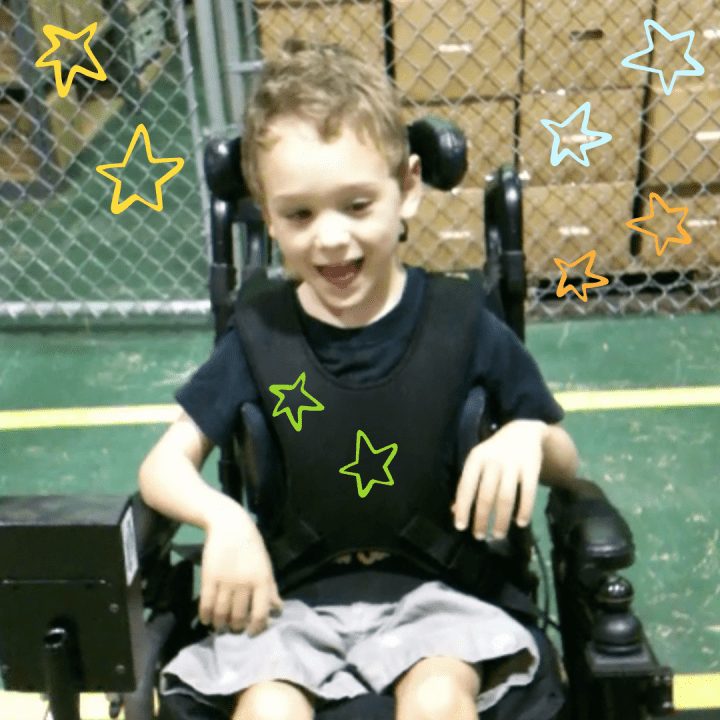 Bower in a power wheelchair wearing a black shirt and tan pants smiling and driving with a head array