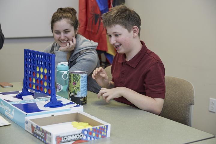 A teenage boy wearing a maroon shirt is playing connect 4 with a teacher with brown hair in a grey sweatshirt smiling.