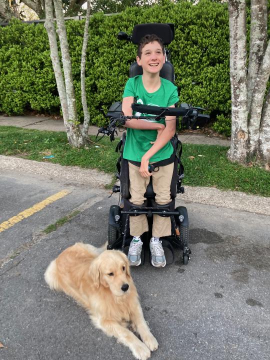 Bower wearing a green shirt and tan pants standing in his wheelchair smiling with his golden retriever everest