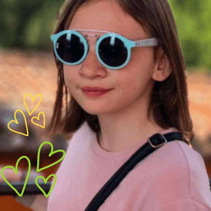 A girl with brown hair is wearing blue framed sunglasses and a pink shirt. She is standing outside with the background of the roof of a historic home