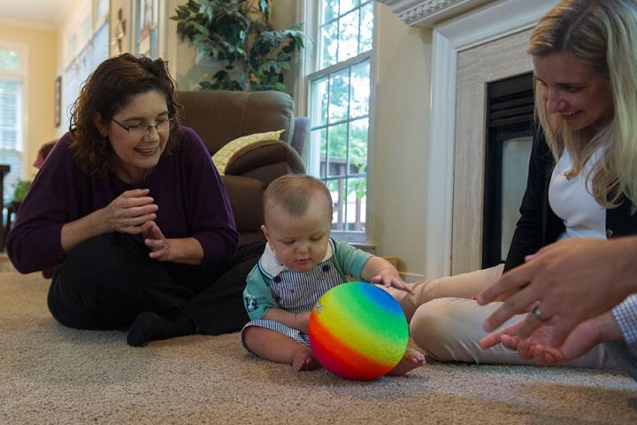 A little baby is sitting on the floor playing with a rainbow colored balloon with his mom, dad and a physical therapist sitting around him smiling and encouraging him.