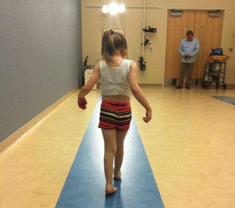 A little girl with brown hair in a pony tail is walking away from the camera on a blue strip on the floor wearing shorts and a t-shirt while being filmed by cameras.
