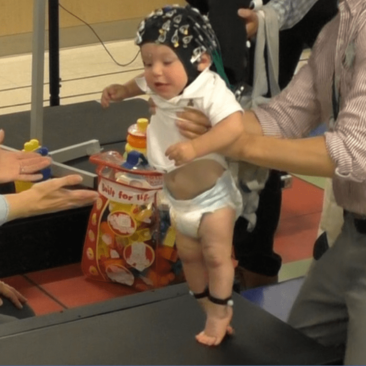 A baby wearing a black eeg cap with a white onesie is being held up by a man while taking a step towards a woman's outstretched hands