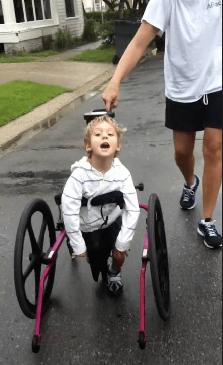 A smiling boy is walking in a gait trainer with the guidance of an adult