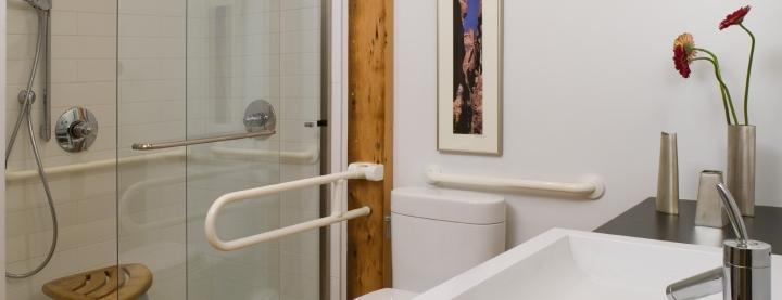 Photo of bathroom looking from sink across toilet to shower. Shower has a grab rail and wooden stool