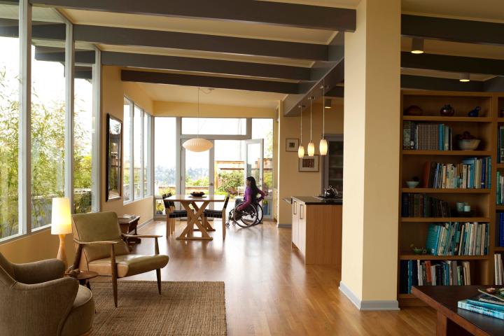 A photo of a modern home with open plan living looking from the lounge room to the dining area and kitchen. There is a young women in a manual wheelchair in the dining room.