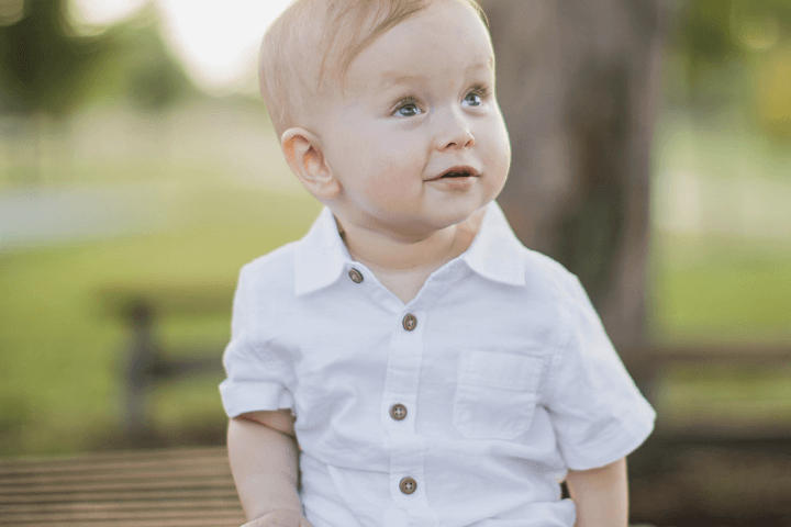 Owen (Age 2) sitting outside under a tree in a white shirt and brown shorts looking up and smiling into the distance