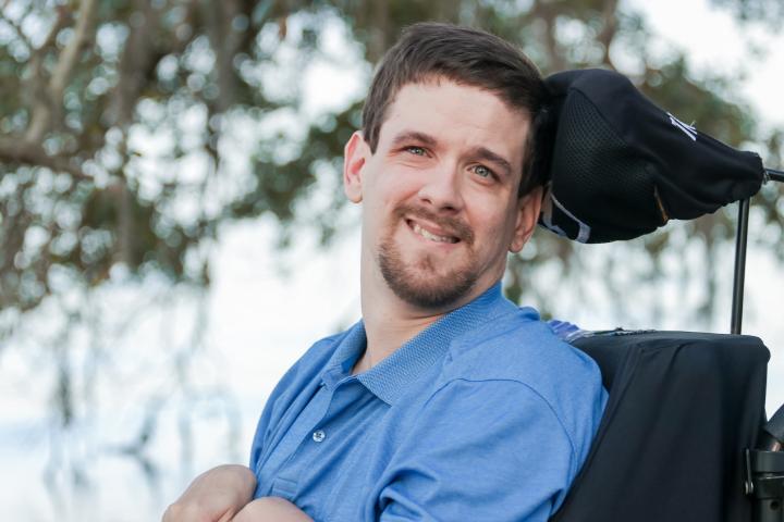 Photo of David Stoner, wearing a blue shirt, sitting in his black wheel chair with a tree in the background.