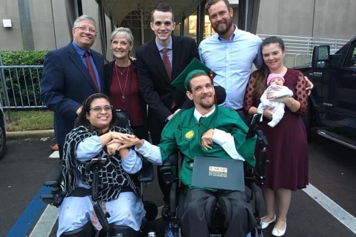 Photograph of a young woman and young man wearing a graduation cap and gown in wheelchairs holding hands with five individuals standing up behind them. One individual is holding an infant. 