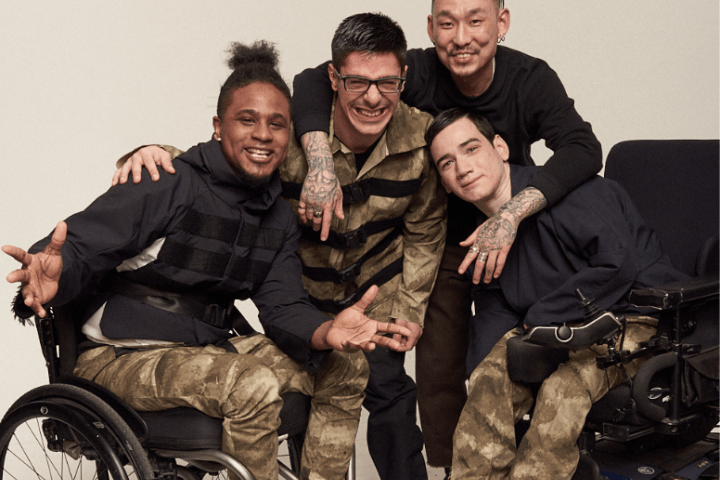 Photo of 4 men: From left to right - Black man, smiling wearing a black turleneck and camo pants seated in a wheelchair next to a white man leaning over smiling with dark hair wearing camo, next to an asian man with dark hair standing, smiling in black turtleneck, next to a white man seated wearing a black turtleneck and camo pants seated in a wheelchair.