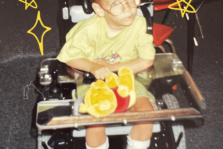 Young David Stoner wearing a yellow shirt and shorts and a pair of glasses in a power wheelchair holding a winnie the pooh teddy bear