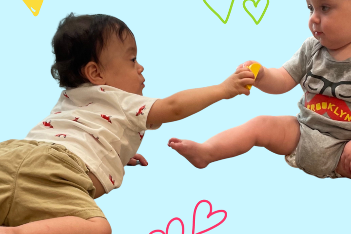Image of a baby with dark hair and tan skin on his belly reaching to give a block to another baby sitting up with a bald head wearing a grey onesie