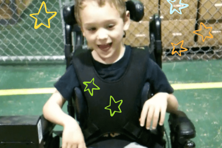 Bower in a power wheelchair wearing a black shirt and tan pants smiling and driving with a head array