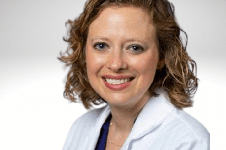  Mary Lauren has curly blond shoulder length hair and is wearing a white lab coat. She is smiling