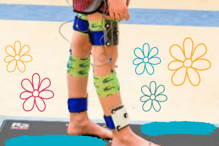 A person's lower body standing up is shown wearing red short and standing on a black mat. The person's legs have sensors around the thighs, knees and ankles with wires attached.