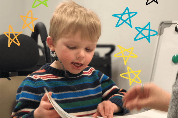 A little boy with blond hair is wear a blue shirt and sitting at table pointing at images on a page.