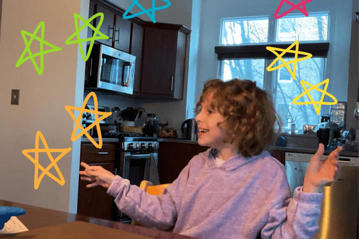 A young girl with medium length curly brown hair is smiling with her arms outstretched while wearing a lilac shirt at a kitchen table in a kitchen of a home.