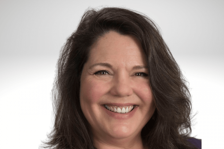 Mary Gannotti has long brown hair and is smiling. She is wear a purple top and a purple beaded necklace.