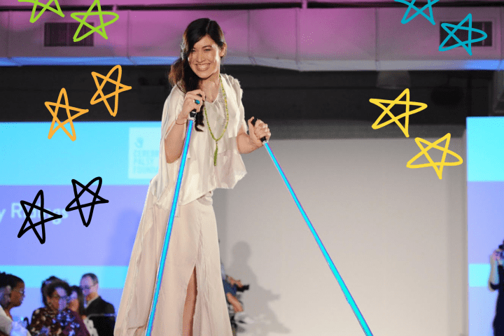 A woman with long brown hair wearing a white flowing dress is on a fashion runway using bright blue poles to walk