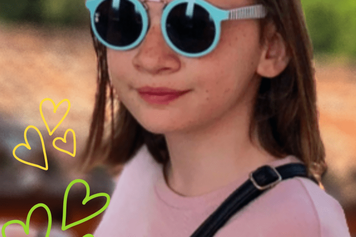 A girl with brown hair is wearing blue framed sunglasses and a pink shirt. She is standing outside with the background of the roof of a historic home