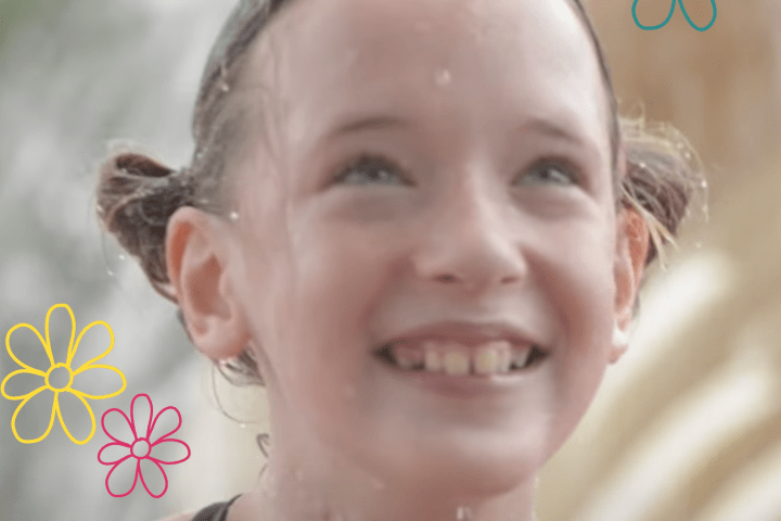 A little girl with blond pigtails and a big smile is in front of a fountain at a water park wearing her swim suit.