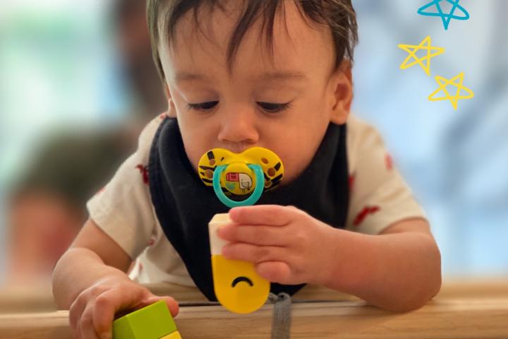 A child with dark hair is sucking on a pacifier while playing with blocks