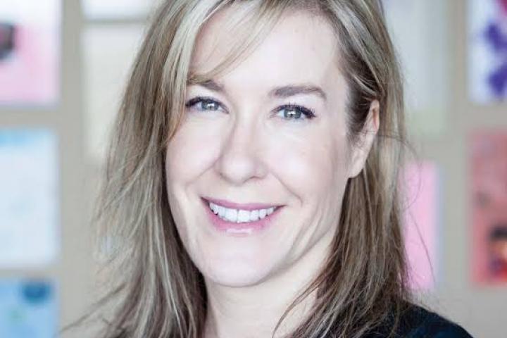 Natalie Maitre has shoulder length sandy blonde hair and blue eyes. She is wearing a blazer and smiling