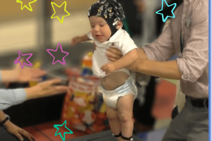 A baby wearing an eeg cap is being held up by a man while he tries to take a step towards a woman with her arms outstretched