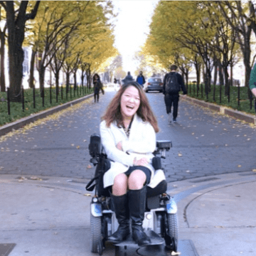 Writer Sarah Kim sitting in her power wheelchair in front of an avenue of trees