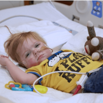 Young child lyign in hospital bed wearing yellow t-shirt and holding a stuffed toy