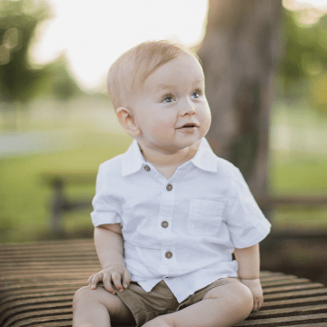 Owen (Age 2) sitting outside under a tree in a white shirt and brown shorts looking up and smiling into the distance