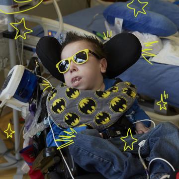 Photo of a young man in a blue shirt, wearing dark sunglasses with yellow rims reclining in a wheelchair with a batman printed neck pillow around his neck. He is in a hospital room and has a feeding tube attached to the wheelchair.  