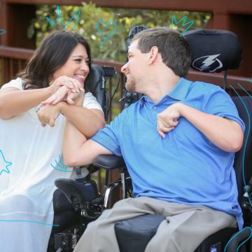 Shelby, a dark haired woman wearing a white dress sitting in a wheelchair next to David who is wear in Blue shirt and khaki pants sitting in his wheelchair.